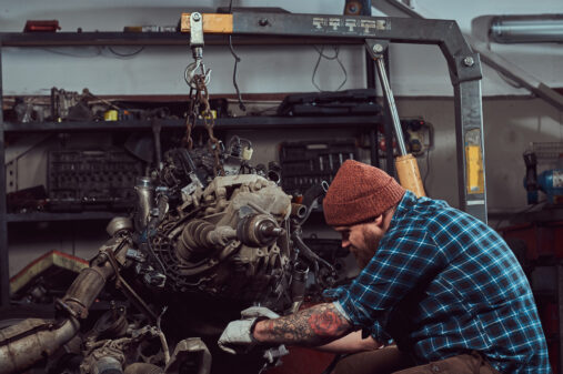 Brutal tattooed bearded mechanic specialist repairs the car engine which is raised on the hydraulic lift in the garage. Service station.